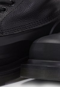 Gros plan sur des chaussures en cuir noir montrant une tige texturée, des œillets noirs et des semelles épaisses en caoutchouc avec des détails rainurés.