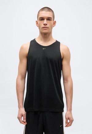 Junger Mann trägt ein schwarzes ärmelloses Nike-Sportshirt und passende schwarze Sportshorts, steht vor einem schlichten hellen Hintergrund.