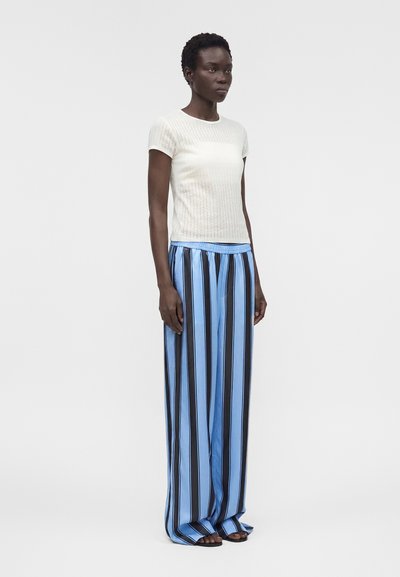 Γυναίκα στέκεται φορώντας ένα λευκό ριμπ κοντομάνικο τοπ και φαρδιά παντελόνια με κάθετες μαύρες και μπλε ρίγες, ξυπόλητη σε απλό φόντο.