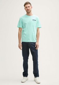 Camiseta de algodón verde menta con un bolsillo en el pecho, combinada con vaqueros de mezclilla azul oscuro y zapatillas blancas. Corte relajado con mangas cortas.