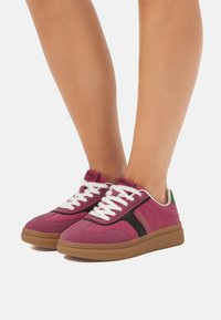 Las zapatillas de ante rosa presentan una franja negra, cordones blancos y una suela de goma marrón con un acento verde en el talón. La textura parece suave y deportiva.