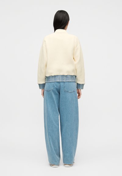 Γυναίκα με μακριά σκούρα μαλλιά που φοράει εκρού σακάκι πάνω από μπλε πουκάμισο με σχέδια και ανοιχτό μπλε τζιν παντελόνα, στέκεται μπροστά σε λευκό φόντο.