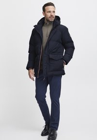 Marineblå jakke med polstring og hætte, med to frontlommer og røde og hvide detaljer. Båret over en lysbrun sweater og marineblå bukser.