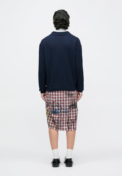 Asmuo stovintis nugara, dėvintis tamsiai mėlyną megztinį, languotą sijoną su abstrakčiais raštais, baltas kojines ir juodus batus, ant paprasto fono.