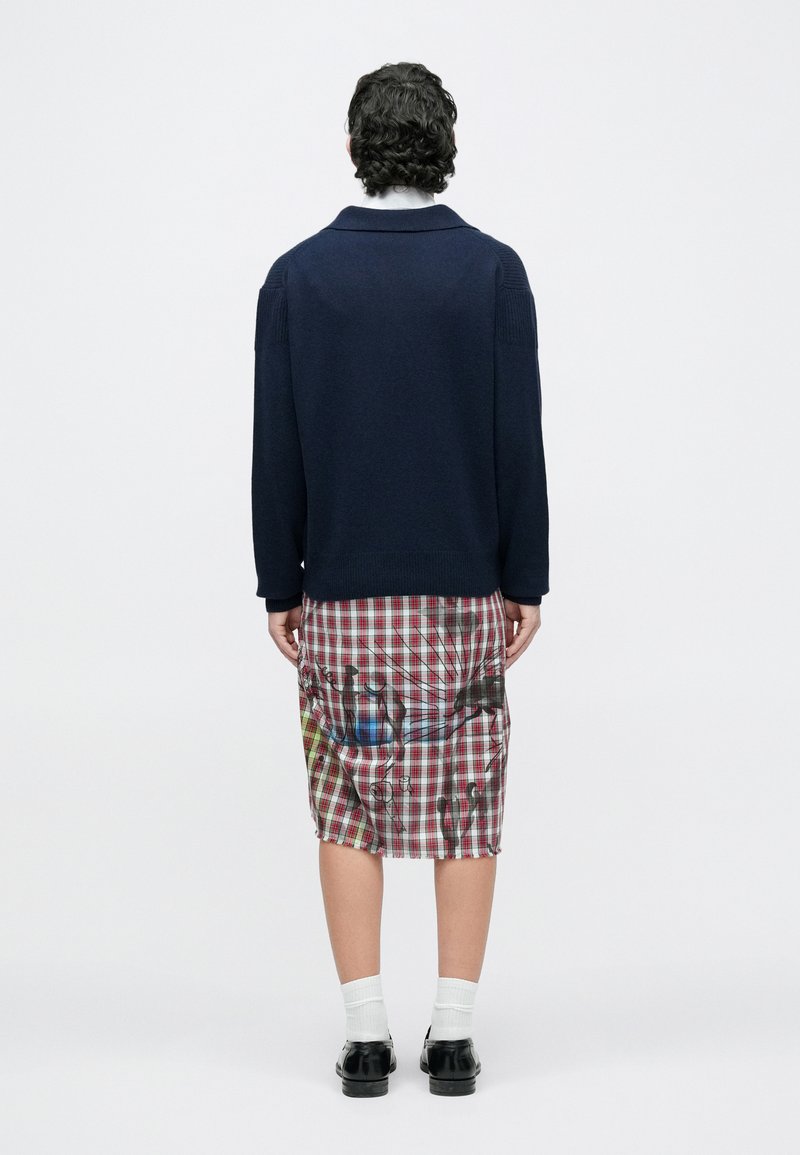 Personne de dos portant un pull bleu marine, une jupe à carreaux avec des motifs abstraits, des chaussettes blanches et des chaussures noires sur un fond uni.