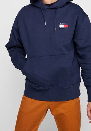 Mężczyzna w granatowej bluzie z kapturem Tommy Jeans z przednią kieszenią i spodniami w spalonym pomarańczowym kolorze, stojący na jednolitym tle.