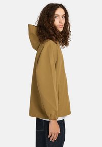 Junge Person mit langen lockigen Haaren, die eine beige Kapuzenjacke und dunkle Jeans trägt und seitlich vor einem schlichten weißen Hintergrund steht.