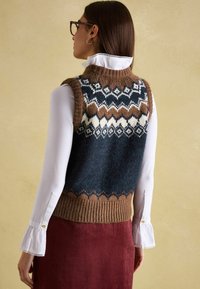 Ärmelloser gestrickter Pullover mit einem geometrischen Muster in Dunkelblau, Braun und Creme, einem gerippten Saum und weicher Textur. Über einem weißen Hemd getragen.