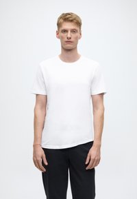 Maglietta bianca a maniche corte in cotone, collo arrotondato, trama liscia, indossata con pantaloni grigio scuro, mostrata su uno sfondo semplice.