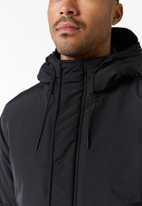 Chaqueta con capucha negra hecha de tejido ligero, con un cuello alto, detalles fruncidos y cordones ajustables para un mejor ajuste.