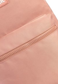 Détail d'un sac à dos en tissu rose avec une poche zippée, une texture lisse et une forme rectangulaire, ornée d'un patch logo en haut à gauche.