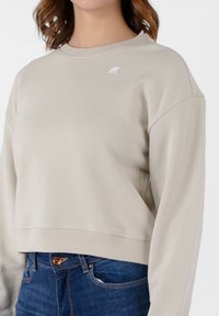 Felpa beige accorciata con texture a costine, colletto rotondo e logo ricamato. Abbinata a jeans di denim blu con bottone di rame.