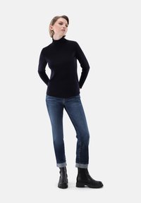 Schwarzer Rollkragenpullover kombiniert mit blauen, umgeschlagenen Jeans und schwarzen Stiefeletten. Der Stoff wirkt glatt; das Outfit hat einen figurbetonten Schnitt.