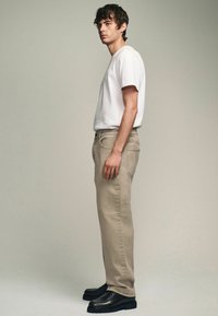 Beige straight-leg pants with side pockets, paired with a fitted white t-shirt. Model wears black chunky shoes. Neutral background.