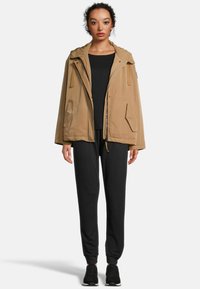 Parka isolée beige avec capuche et fermeture éclair à l'avant, comportant deux poches latérales ; portée sur un haut noir et un jogging noir. Baskets noires aux pieds.