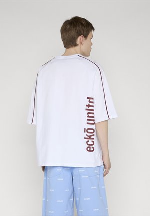 Mladý muž tvárou do boku nosí nadrozmerné biele tričko s nápisom "eckō unltd" a svetlomodré šortky s opakujúcim sa vzorom značkového loga.
