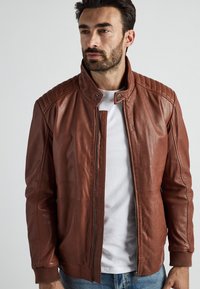 Ruskea nahkatakki, jossa on vetoketju edessä, seisova kaulus ja quiltatut olkapäät. Ominaisuuksiin kuuluu teksturoitu materiaali ja ribatut hihansuut.