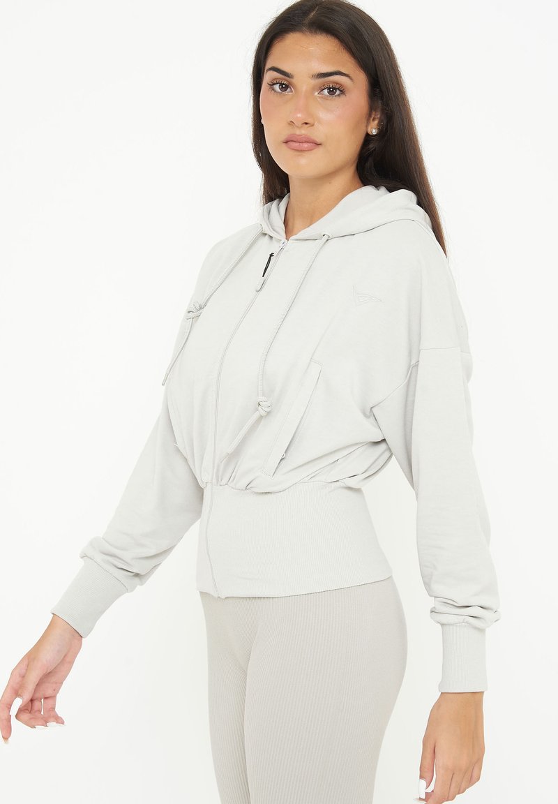 Hoodie corto grigio chiaro con lacci, zip frontale e orlo a costine. Realizzato in tessuto morbido con una texture liscia.
