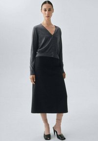 Grauer, langärmeliger Cardigan mit V-Ausschnitt und Knöpfen, kombiniert mit einem schwarzen, knöchellangen Rock. Das Model trägt rosa gerippte Socken und schwarze hohe Schuhe.