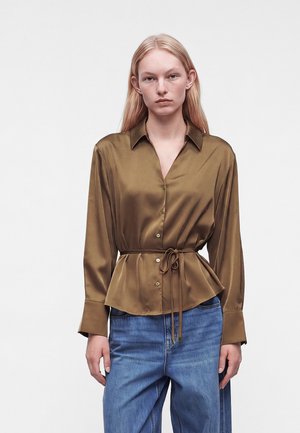Femme portant un chemisier en satin marron boutonné avec une ceinture nouée à la taille et un jean bleu, debout devant un fond clair uni.