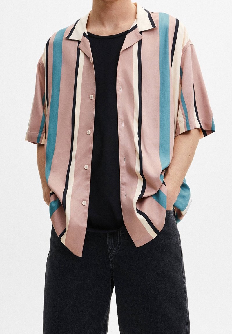 Homme portant une chemise ample à manches courtes boutonnée avec des rayures verticales rose, beige, bleu-vert et noir, par-dessus un t-shirt noir et un pantalon foncé.