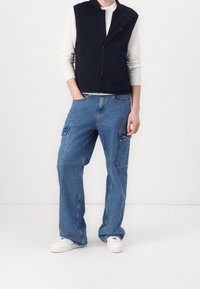 Osoba nosząca granatową kurtkę na zamek błyskawiczny over białą koszulę z długim rękawem, luźne niebieskie spodnie cargo oraz białe trampki, stojąca z jedną ręką w kieszeni.