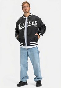 Casaco bomber preto com texto branco, punhos listrados e ténis pretos, combinado com jeans baggy azul claro numa pose de pé.