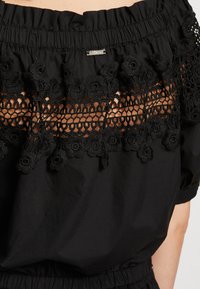 Top nero scollato sulle spalle con dettagli in pizzo intricati e motivi floreali in un tessuto testurizzato. Vita elastica e accento di hardware sottile.