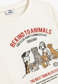 Sudadera crema con un gráfico de varios perros, con texto en rojo y negro promoviendo la adopción de animales. Textura suave, cuello acanalado.