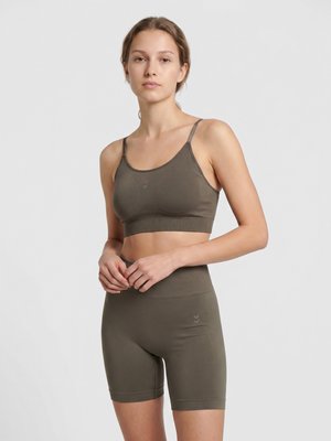 Ung kvinde stående med foldede hænder iført olivengrøn sports-bh og højtaljede cykelshorts mod en ensartet lys baggrund.