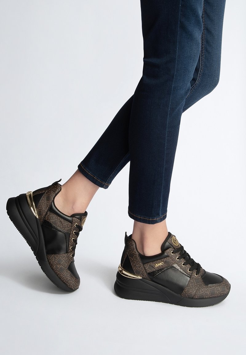 Pieds portant des baskets noires et marron à motifs avec des accents dorés, assorties à un jean skinny bleu foncé sur fond blanc.