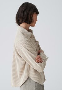 Light beige corduroy shirt with a textured ribbed pattern, featuring a collar, button details, and slightly curved hem.