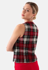 Geruite mouwloze vest in rood, zwart en witte stof, met een aansluitend ontwerp en zonder sluitingen, gecombineerd met rode broeken.