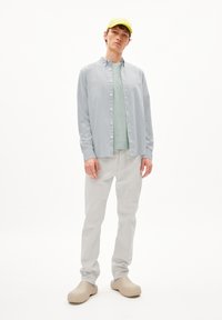 Lichtgrijs button-up shirt over een mintgroene t-shirt, gecombineerd met lichtgrijze broek en gestructureerde beige klompen, met een neongroene pet.