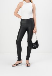 Leggings taille haute en simili cuir noir, associés à un débardeur blanc à côtes et un sac à main noir avec des lettres argentées, sur fond neutre.
