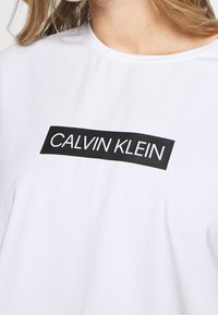 Biała bawełniana koszulka z czarnym prostokątnym logo z napisem "CALVIN KLEIN" w białym kolorze. Okrągły dekolt i krótkie rękawy.
