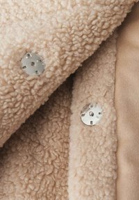 Tissu beige duveteux avec une apparence texturée, comprenant des boutons-pression en argent et une doublure lisse.