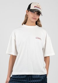 Femme portant un t-shirt blanc oversize avec un petit texte rouge et une casquette beige-noire avec un logo rouge et bleu, debout devant un fond uni.