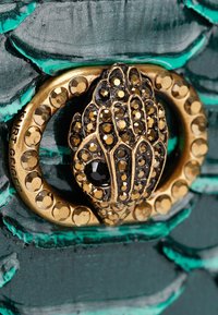 Goudkleurige sluiting met een ingewikkeld handontwerp, versierd met kleine edelstenen op een teal-geweven leren achtergrond.