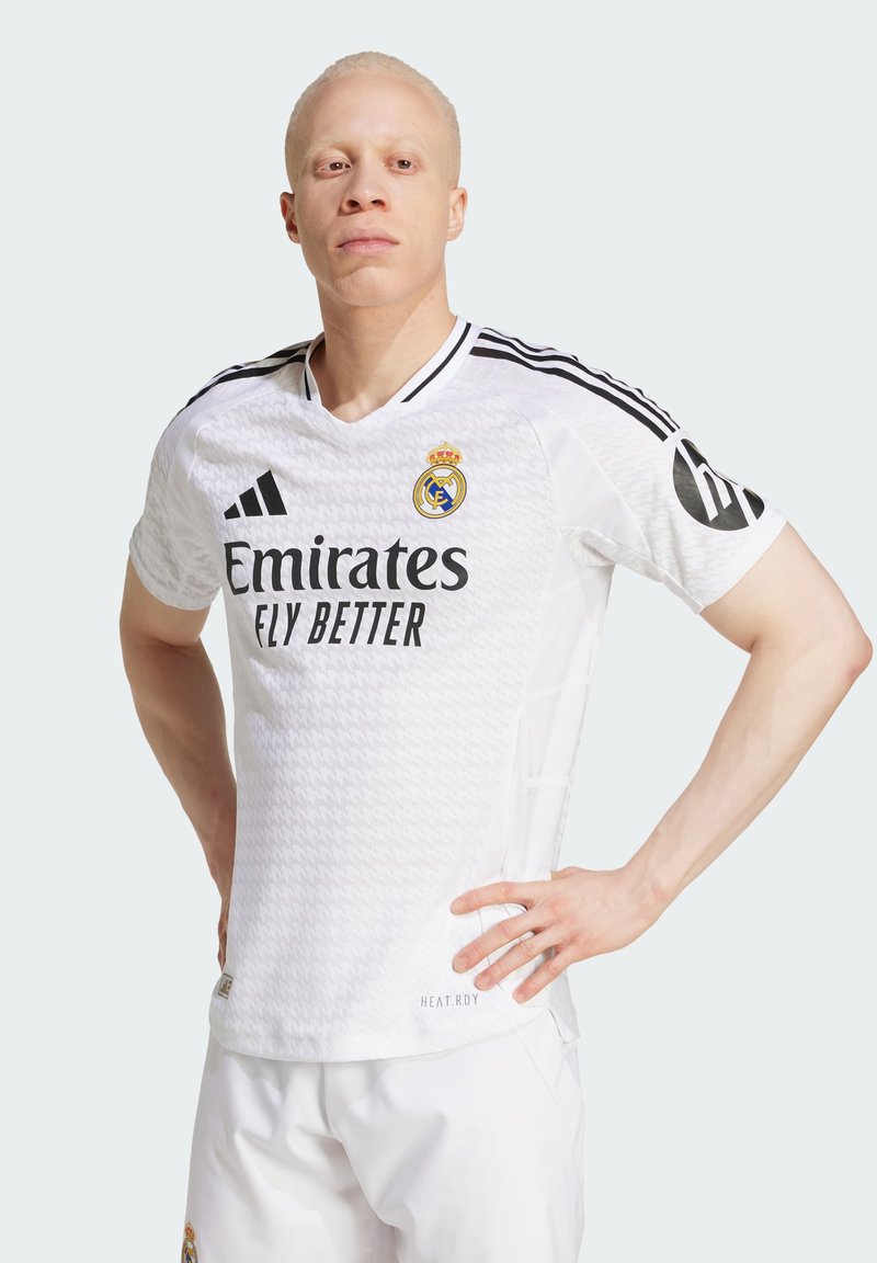 Camiseta de fútbol blanca del Real Madrid con un patrón texturizado, acentos negros y diseño de cuello en V. Presenta el logo "Emirates ELY BETTER" y las rayas de adidas.