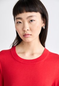 Κόκκινο πλέ knitted πουλόβερ με στρογγυλή λαιμόκοψη. Το μοντέλο έχει ίσια μαύρα μαλλιά και φοράει ένα ασημένιο σκουλαρίκι βολβού. Η υφή φαίνεται μαλακή και λεία.