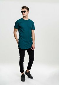 Tirkizna pamučna majica s okruglim izrezom, uparena s crnim teksturiranim slim-fit trapericama i crnim tenisicama. Model nosi sunčane naočale i stoji ležerno.