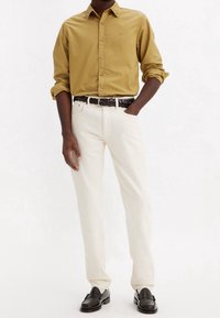 Camisa abotonada en mostaza, combinada con jeans de mezclilla blancos. Un cinturón de cuero negro trenzado y unos mocasines negros completan el look.