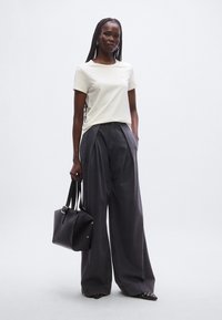 Kremowy t-shirt z krótkim rękawem w połączeniu z ciemnymi, szerokimi spodniami z wysokim stanem. Modelka trzyma czarną skórzaną torebkę. Prosty design, minimalne akcenty.