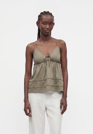 Donna di colore con capelli intrecciati che indossa un top verde oliva senza maniche con nodo frontale e pantaloni bianchi a gamba larga, in piedi davanti a uno sfondo semplice.