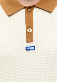 Gros plan d’un polo beige texturé avec col et patte marron, deux boutons, et un écusson logo bleu "HUGO" sous les boutons.