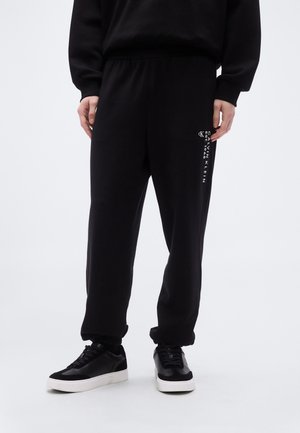 Črne trenirke z ohlapnim krojem, elastičnim pasom in belim tiskom logotipa Calvin Klein na strani. Ujemajo s črnimi supergami.