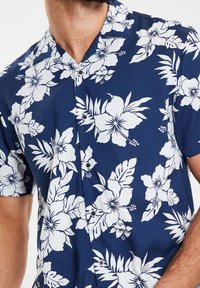 Lyhythihainen paita, jossa on laivastonsininen kangas, suuret valkoiset kukkakuviot ja vihreät lehdet, napitettu klassisella kauluksella.