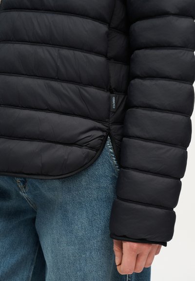 Μανίκι και πλευρό μαύρου μπουφάν με σχέδιο καπιτονέ πάνω από μπλε τζιν, δείχνοντας την ετικέτα «CLOSED» στη ραφή του μπουφάν.