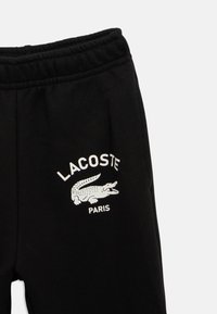 Sorte sweatpants med elastisk talje har et hvidt trykt Lacoste-logo og krokodile-design på venstre lår. Blød tekstur.